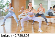Group of positive sporty women of different ages in activewear performing squats before dance class with trainer in hall. Стоковое фото, фотограф Яков Филимонов / Фотобанк Лори