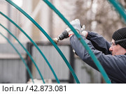 A man works with an electric grinder to clean rust from metal iron structures. Rust removal using an electric iron brush. Hands in work gloves are holding a power tool. Стоковое фото, фотограф Photojuli / Фотобанк Лори