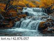 Picturesque waterfall in the forest in autumn. The water rushes forcefully over multi-level rocky rapids, creating a striking contrast with the warm colors of the autumn foliage surrounding the pond. The leaves of the trees, painted in golden and copper tones, glow against the background of dark blue water and stones, which adds peace and harmony to the scene. Стоковое AI-изображение, AI-художник Анна Кривицкая / Фотобанк Лори