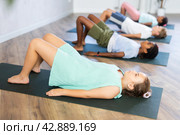 Children doing yoga with group in studio, lying on mat with knee pressed to chest with hands, performing hamstring and low back stretching Ardha Pawanmuktasana. Стоковое фото, фотограф Яков Филимонов / Фотобанк Лори