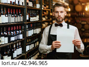 A professional sommelier stands in the wine cellar and holds a white blank sheet. Стоковое AI-изображение, AI-художник Ярославский Максим / Фотобанк Лори