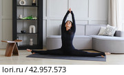 A woman does yoga at home, performs the exercise Samakonasana, transverse twine, trains in a black sports overalls on the mat, healthy lifestyle concept. Стоковое фото, фотограф Сергей Чайко / Фотобанк Лори