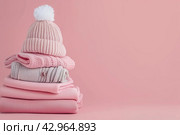 Stack of baby knitted clothes on a table. Set of newborn child rompers, hat and jumper. Fashion infant accessories isolated pastel pink background, empty space. Kid's toys and clothes. Стоковое AI-изображение, AI-художник Марина Демидюк / Фотобанк Лори