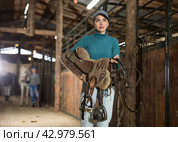 Young horsewoman walking in stable with saddle in hands. Стоковое фото, фотограф Яков Филимонов / Фотобанк Лори