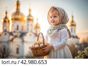 Happy bright Easter. Christianity. Portrait of a three-year-old girl in a Russian folk dress and shawl with a wicker basket with Easter eggs in her hands against the background of an Orthodox church. Стоковое AI-изображение, AI-художник Photojuli / Фотобанк Лори