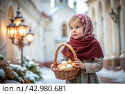 Happy bright Easter. Christianity. Portrait of a three-year-old girl in a Russian folk dress and shawl with a wicker basket with Easter eggs in her hands against the background of an Orthodox church. Стоковое AI-изображение, AI-художник Photojuli / Фотобанк Лори