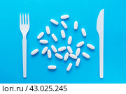 White oval pills and knife with a fork on a blue background. Top view. Uncontrolled use of drugs in large quantities. Стоковое фото, фотограф Елена Руй / Фотобанк Лори