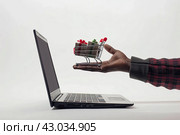 A man's hand holds a small shopping cart full of goods against the background of a laptop. The concept of online shopping. Стоковое AI-изображение, AI-художник Ярославский Максим / Фотобанк Лори