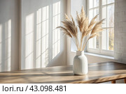 A modern white ceramic vase with dried grass on a wooden table. Scandinavian interior. An empty white wall with a shadow from the window. Стоковое AI-изображение, AI-художник Photojuli / Фотобанк Лори