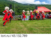Adygea, RUSSIA - JULY 25 2015: A group of Circassian boys and girls in beautiful red national costumes and white sheepskin hats dance on a green meadow against the background of mountains. Редакционное фото, фотограф Андрей С / Фотобанк Лори