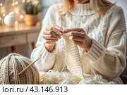 Close-up. A woman with balls of yarn knits a warm sweater in white and beige tones in her hands. Needlework, knitting. Winter season, small business, handmade clothes. Стоковое AI-изображение, AI-художник Photojuli / Фотобанк Лори