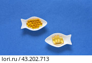 Dietary supplements or nutrition additions with fish oil, vitamin E and omega acids. Concept of preventing heart disease using fish oil. Flat lay of gelatin capsules on fish-shaped plates on blue background. Стоковое фото, фотограф Виктория Катьянова / Фотобанк Лори