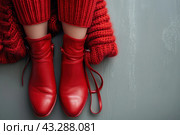 The stylish and cozy image shows a woman's legs wearing red leather boots and a thick red knitted sweater. The woman sits on the gray floor, and her image conveys a feeling of warmth and comfort. Red boots and a sweater create a pop of color that contrasts with the cool gray background. The shoes look high quality and fashionable, and the sweater adds coziness and completeness to the look. Стоковое AI-изображение, AI-художник Анна Кривицкая / Фотобанк Лори