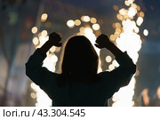 Silhouette of young excited woman during street rock concert. Woman waves her arms to the beat of the music. She dances. Huge sparkler candles are burning. Bright night life of the big city. Back view. Стоковое фото, фотограф YuryKara / Фотобанк Лори