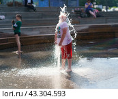 Boy washing hif face by cold water of fountain in the hot summer day in the big city. Fresh water splashes. He is wet. Concept of the leisure time, freshness and happiness. Frontal view (2021) Редакционное фото, фотограф YuryKara / Фотобанк Лори