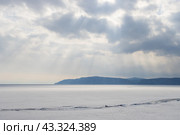Озеро Байкал, Иркутская область, Россия. Лучи солнца проходят через промежутки в облаках и освещают покрытое льдом озеро Байкал. Вдали гористый берег. Красивый пейзаж. Стоковое фото, фотограф Андрей Степанов / Фотобанк Лори