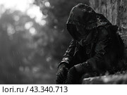 Man sitting by the wall in the rain. The picture is made in black and white, which enhances the feeling of sadness and depression. The man is wearing a dark jacket with a hood that hides his face and gloves. Raindrops are visible on his clothes, creating an atmosphere of cold and hopelessness. The man is bowed, head and hands bowed, his posture expresses deep sadness and loneliness. Стоковое AI-изображение, AI-художник Анна Кривицкая / Фотобанк Лори