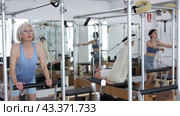 Svelte elderly woman performing set of pilates exercises on reformer during group workout. Active lifestyle and wellness concept of seniors. Стоковое видео, видеограф Яков Филимонов / Фотобанк Лори