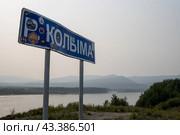 Придорожный информационный знак с названием реки Колыма. Знак на федеральной автомобильной дороге «Колыма» (Колымская трасса). На знаке много наклеек туристических экспедиций. Вдали река Колыма и горы в дымке. Магаданская область, Россия. Автомобильное путешествие по Сибири и Дальнему Востоку России. (2023 год). Редакционное фото, фотограф Андрей Степанов / Фотобанк Лори