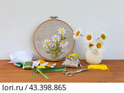 Handmade embroidery with satin ribbons of daisies in hoop, bouquet of fresh daisy flowers in vase on wooden table. Nearby is set of colorful satin ribbons, needles and scissors for work. DIY concept. Стоковое фото, фотограф Виктория Катьянова / Фотобанк Лори