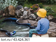 A 5-year-old boy in a yellow panama hat launches a radio-controlled boat in a pond. Children's fun and street games. Child's leisure time. Стоковое фото, фотограф Photojuli / Фотобанк Лори