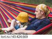 Summer holidays for children in the fresh air. A cute little boy with his father is having fun on a hammock in the backyard. A child and a dad in a hammock. Стоковое фото, фотограф Photojuli / Фотобанк Лори