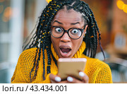 A young black woman with glasses and a surprised expression keeps a mobile phone at home. Стоковое AI-изображение, AI-художник Ярославский Максим / Фотобанк Лори