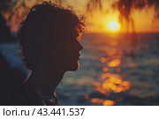 Silhouette of a young man against the backdrop of sunset by the sea. The sun has almost disappeared below the horizon, painting the sky and sea in warm orange and golden tones. Tree branches create a natural frame for the image. The silhouette of a person stands out against the backdrop of a bright sunset, creating an atmosphere of thoughtfulness and peace. The glare of sunlight reflects on the water, adding picturesqueness and magic to the picture. Стоковое AI-изображение, AI-художник Анна Кривицкая / Фотобанк Лори