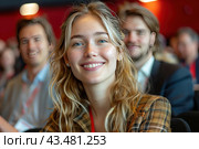Smiling young people sitting in the audience. In the foreground is a girl with long blond hair, wearing a checkered jacket. She smiles widely and looks happy and content. In the background, other people are visible, also smiling and, apparently, attentively listening to some event. The overall atmosphere of the image is positive and friendly. Стоковое AI-изображение, AI-художник Анна Кривицкая / Фотобанк Лори