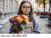 A pretty little girl with glasses and a bouquet of autumn flowers goes to school for the first time. A child with a school bag, a book and flowers. A child on a city street. Back to school. Стоковое AI-изображение, AI-художник Photojuli / Фотобанк Лори