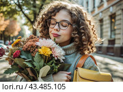 A cute little curly-haired girl with glasses and a bouquet of autumn flowers goes to school for the first time. A child with a school bag, a book and flowers. A child on a city street. Back to school. Стоковое AI-изображение, AI-художник Photojuli / Фотобанк Лори