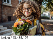 A cute little curly-haired girl with glasses and a bouquet of autumn flowers goes to school for the first time. A child with a school bag, a book and flowers. A child on a city street. Back to school. Стоковое AI-изображение, AI-художник Photojuli / Фотобанк Лори