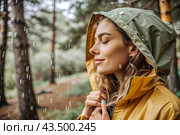 Close-up. A happy young woman in a raincoat enjoys the rain on the background of a green forest, the concept of enjoying a natural leisurely life, environment, lifestyle. Стоковое AI-изображение, AI-художник Photojuli / Фотобанк Лори