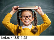 A pretty girl with glasses and books on her head. The child goes to school for the first time. A child in the classroom on the background of a blackboard. Back to school. Стоковое AI-изображение, AI-художник Photojuli / Фотобанк Лори