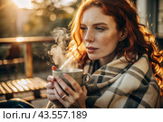 Autumn evening. A red-haired, beautiful woman wrapped in a plaid with a mug of hot coffee on a beautiful patio in the rays of the setting sun. The girl inhales the aroma of the drink. Romance, comfort and harmony. Стоковое AI-изображение, AI-художник Photojuli / Фотобанк Лори
