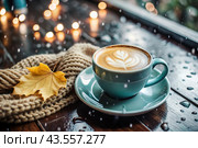 A seasonal drink. Delicious hot cappuccino in a mug with a saucer and a knitted snood, a scarf on a wooden table in the interior of the cafe. It's a rainy autumn day. Стоковое AI-изображение, AI-художник Photojuli / Фотобанк Лори