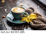 A seasonal drink. Delicious hot cappuccino in a mug with a saucer and a knitted snood, a scarf on a wooden table in the interior of the cafe. It's a rainy autumn day. Стоковое AI-изображение, AI-художник Photojuli / Фотобанк Лори