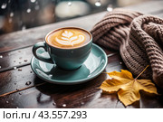 A seasonal drink. Delicious hot cappuccino in a mug with a saucer and a knitted snood, a scarf on a wooden table in the interior of the cafe. It's a rainy autumn day. Стоковое AI-изображение, AI-художник Photojuli / Фотобанк Лори
