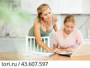 Mother looks at laptop and prompts and helps teen girl to write in notebook to complete task. Стоковое фото, фотограф Яков Филимонов / Фотобанк Лори