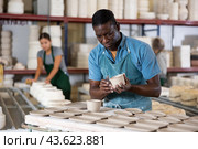 Portrait of african american worker cutting burrs off freshly made earthenware cups and plates with knife. Стоковое фото, фотограф Яков Филимонов / Фотобанк Лори