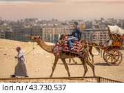CAIRO, EGYPT - MAY 18 2021: Bedouin camel driver rides an Asian tourist from China on a camel, on the Giza plateau. Asian tourist travel to Egypt. Редакционное фото, фотограф Павел Муравьев / Фотобанк Лори