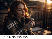 A red-haired, beautiful woman wrapped in a plaid with a mug of hot coffee on a beautiful patio in the rays of the setting sun. The girl inhales the aroma of the drink. Comfort and harmony. Стоковое AI-изображение, AI-художник Photojuli / Фотобанк Лори