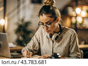 A young female freelance businessman in glasses and headphones is working on a laptop or shopping in an online store. The woman is writing something in a notebook. Стоковое AI-изображение, AI-художник Photojuli / Фотобанк Лори