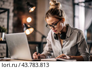 A young female freelance businessman in glasses and headphones is working on a laptop or shopping in an online store. The woman is writing something in a notebook. Стоковое AI-изображение, AI-художник Photojuli / Фотобанк Лори
