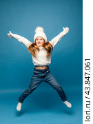 Child girl in a white sweatshirt and knitted hat with pompom jumping on a blue background. The concept of children's emotions, joy and delight. Advertising clothing and sales. Ski resort vacation. Стоковое фото, фотограф Ирина Михайличенко / Фотобанк Лори