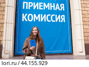 Красивая девушка-подросток держит в руках аттестат об окончании российской школы, стоит возле института, перед вывеской с надписью «Приемная комиссия». Молодая женщина поступает в университет. Стоковое фото, фотограф Яна Проничева / Фотобанк Лори
