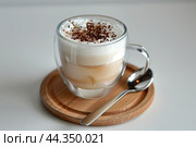 Cafe latte macchiato layered coffee in a see through coffee cup. Wooden tray with milk coffee in a transparent trendy glass mug, spoon on a white desk. Hot cappuccino coffee on table. Space for text. Стоковое AI-изображение, AI-художник Марина Демидюк / Фотобанк Лори