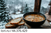 A warm bowl of hearty soup accompanied by bread, set against a snowy mountain backdrop on a chilly winter day. Стоковое AI-изображение, AI-художник Сергей Толмачёв / Фотобанк Лори
