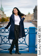 Charming thoughtful fashionably dressed woman with long dark hair travels through Europe, standing in city center of St. Petersburg. A beautiful girl wanders alone through autumn streets. Стоковое фото, фотограф Natalia Gdovskaia / Фотобанк Лори