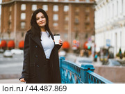 Beautiful serious smart brunette girl holding cup of coffee in hands goes walking down street in center on blue bridge. Charming thoughtful woman with long hair wanders alone, immersed in thoughts. Стоковое фото, фотограф Natalia Gdovskaia / Фотобанк Лори